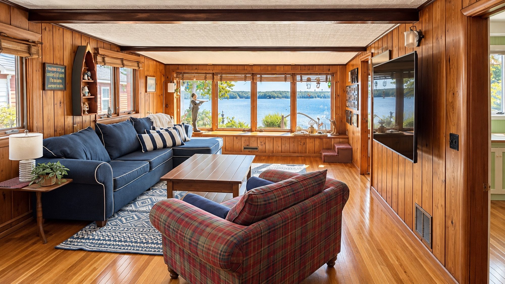 Lakefront living room of a Raymond, New Hampshire home after a J&B Pro Cleaning visit: knotty pine walls, a navy sectional, a plaid armchair and polished wood floors, with the lake through the window wall
